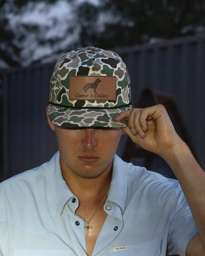 A man wearing a duck camouflage SnapBack cap with a brown leather patch that reads 'Cameo & Collar' and a dog silhouette, holding the brim down over his eyes. He is wearing a light blue button-down shirt.